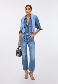 Cămașă din denim, pullover albastru pe umeri, blugi albaștri deschiși, poșetă neagră, pantofi cu toc argintii cu model de piele de șarpe, coliere suprapuse.