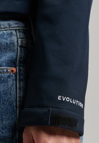 Mörkblå jackärm med kardborreband. Utrustad med vit broderad "EVOLUTION." Slät, matt textur, närbild av tyg och söm.