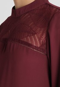Burgundy-blus med hög hals, detaljer av intrikat spets över bröstet och slät tyg som ger en flytande silhuett.