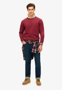 Rød sweatshirt med rund hals, lys skjorte med krave under, blå jeans og brune sko. Ternet flannel skjorte bundet omkring livet.