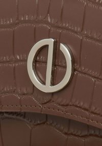 Brązowa skórzana torebka z tłoczeniem w kształcie krokodyla z metalicznym okrągłym detalem logo. Gładka tekstura, widoczne szwy boczne i błyszczące wykończenie.