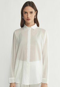 Blusa bianca a maniche lunghe con colletto a camicia, dal tessuto leggermente trasparente e orlo dritto. Presenta un design semplice e pulito con minimi accenti.