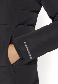 Chaqueta acolchada negra con bolsillo con cremallera, diseño de paneles cosidos y puños elásticos, con el logo de "Calvin Klein Jeans" en la manga.