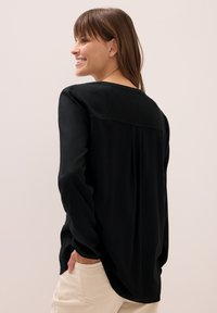 Schwarze Bluse mit langen Ärmeln und lockerem Schnitt, mit einer gefältelten Rückseite, weichem Material und rundem Halsausschnitt. Kombiniert mit hellen Hosen.
