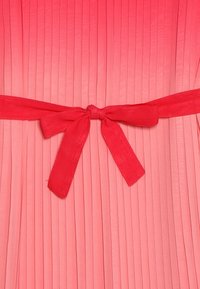 Pilkullinen mekko, jossa on vaaleanpunaisen sävyjen liukuväri, leveä punainen satiinivyö solmittuna rusetille vyötäröllä. Kuvioitu kangas pystysuunnassa olevilla laskoksilla.