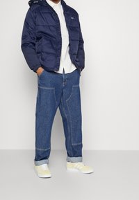 Marinblå puffjacka med huva, vit skjorta under, lösa blå denimbyxor med kontrasterande sömmar och gula sneakers med vita ränder.