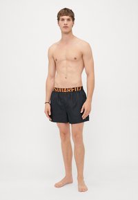 Junger Mann, der barfuß in schwarzen Badeshorts mit orangefarbener Taille steht, vor einem schlichten hellen Hintergrund.