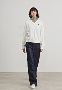 Witte v-hals sweatshirt met geribbelde manchetten, gecombineerd met hooggetailleerde donkerblauwe spijkerbroek en witte sneakers. Model staat in een neutrale omgeving.