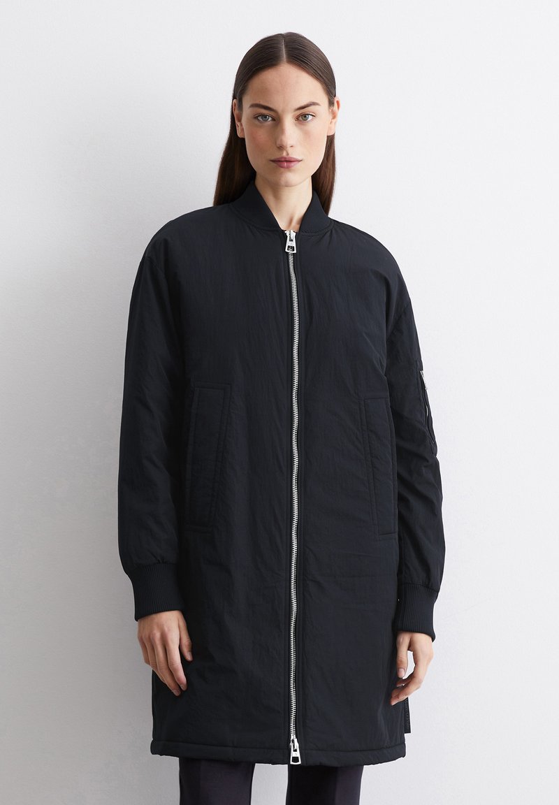 Veste bomber noire avec fermeture éclair à l'avant, poignets côtelés, poches latérales et texture lisse. Présente un design minimaliste et une coupe ajustée.