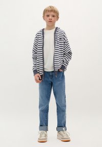 Gestreepte hoodie in marineblauw en wit, lichte stof, grijze accenten, gedragen over een wit T-shirt. Lichtblauwe denim jeans met opgerolde mouwen, neutrale sneakers.