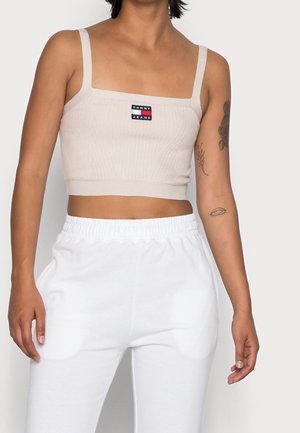 Osoba w beżowym krótkim topie z logo Tommy Jeans i białych spodniach z elastycznym pasem, pokazująca tatuaże na rękach na jasnym tle.
