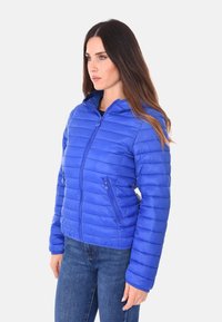 Giacca a puffo blu con zip frontale, motivo a colonne orizzontali e due tasche laterali. Design aderente con cappuccio e texture liscia.