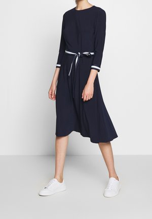 Kobieta ubrana w granatową sukienkę sięgającą kolan z rękawami do łokcia, mankietami z białymi paskami, przewiązanym paskiem w talii i białymi trampkami.