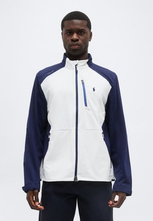 Mężczyzna ubrany w białą i granatową kurtkę zapinaną na suwak z małym logo na klatce piersiowej, stojący na tle jednolitego, jasnego tła.