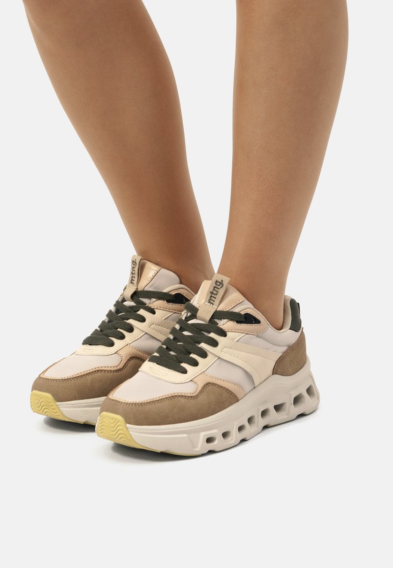 Chunky Sneaker mit beigem Synthetik-Obermaterial, olivgrünen Schnürsenkeln, Wildlederakzenten und einer dicken weißen Gummisohle mit Ausschnitten.