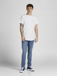 Weißes T-Shirt mit Rundhalsausschnitt und kleinem Logo, kombiniert mit hellblauen Slim-Fit-Jeans und dunkelblauen Sneakern mit weißen Sohlen. Einheitlicher Hintergrund.