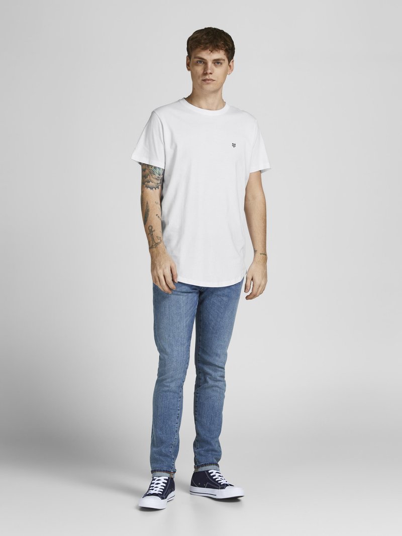 Weißes T-Shirt mit Rundhalsausschnitt und kleinem Logo, kombiniert mit hellblauen Slim-Fit-Jeans und dunkelblauen Sneakern mit weißen Sohlen. Einheitlicher Hintergrund.
