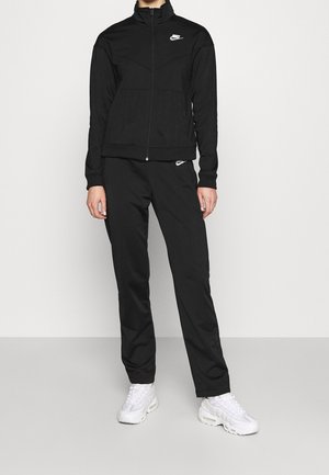Personne portant une veste de survêtement Nike noire et un pantalon assorti, avec des baskets Nike blanches, debout devant un fond clair uni.