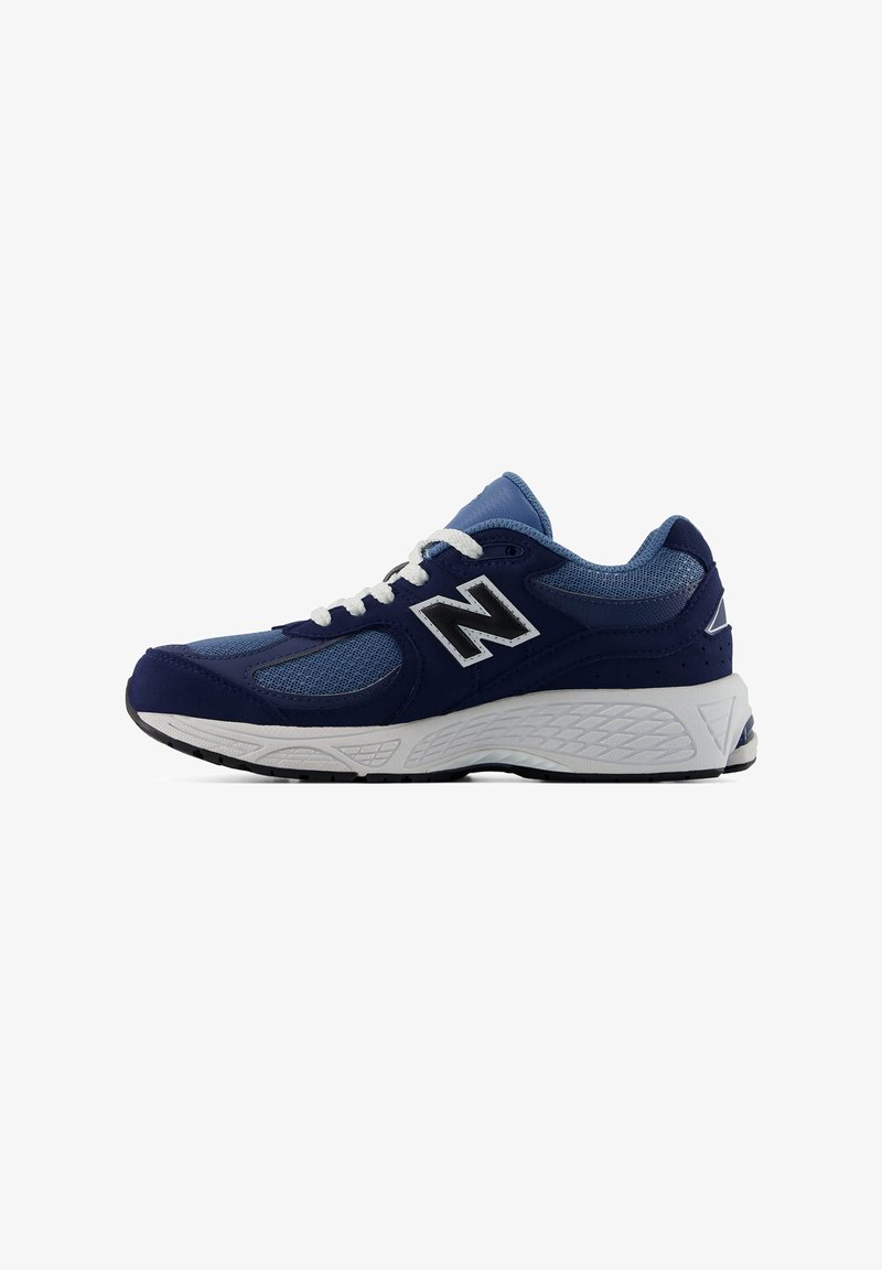 Τζιν και μπλε αθλητικό παπούτσι New Balance με λευκά κορδόνια και παχύ λευκό υφασμένο πέλμα, εμφανιζόμενο από το πλάι σε λευκό φόντο.