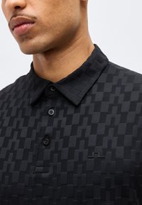 Κοντινό πλάνο ενός άνδρα που φοράει μαύρο πολο με κουμπιά και γεωμετρικό υφασμάτινο μοτίβο με πιεσμένο γιακά.