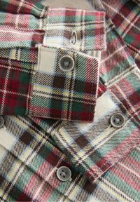 Gros plan sur un tissu à carreaux plié aux couleurs rouge, vert, marron et crème, montrant des boutons gris et des boutonnières sur les poignets de chemise.