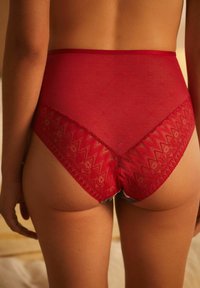 Hochgeschnittene rote Unterwäsche mit einem transparenten Netzrücken und aufwendigen Spitzenverzierungen, die ein Zickzackmuster an den Kanten aufweisen.