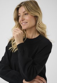 Maglione nero con colletto rotondo e maniche lunghe, caratterizzato da polsini scallopati. Tessuto liscio e vestibilità comoda. Il modello sorride.