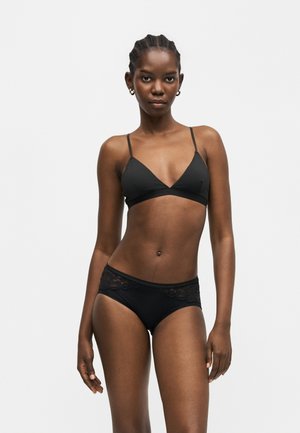 Femme aux cheveux tressés portant un soutien-gorge triangle noir et des sous-vêtements assortis avec des détails en dentelle, debout devant un fond gris clair uni.