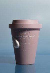 Różowy cylindryczny pojemnik z pokrywką, z kroplami wilgoci i białą teksturą kremu po boku. Etykieta zawiera nazwę produktu oraz szczegóły.