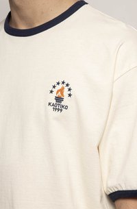Cream t-shirt with navy ribbed neckline. Features embroidered logo with flame and stars in navy and orange. Short, loose sleeves.