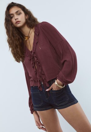 Una giovane donna con lunghi capelli ricci indossa una blusa bordeaux con volant, pantaloncini di denim scuro, una collana d'oro a forma di fiore e braccialetti d'oro su uno sfondo chiaro.