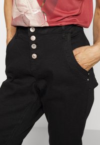 Calças pretas de cintura alta com cinco botões decorativos, bolsos laterais e um tecido texturizado, apresentando detalhes metálicos discretos.