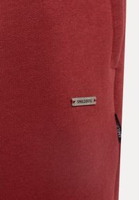 Röd bomullstyg med en jämn yta, utrustad med en liten metallisk logotypetikett med inskriptionen "SMILODOX." Enkel design, utan mönster.