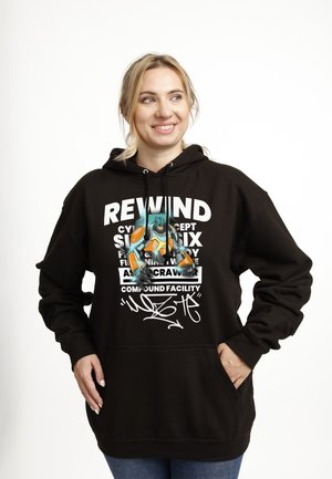 Kvinde i sort hættetrøje med farverigt robotmotiv og hvid tekst, stående med hænderne i lommerne, smilende og kiggende til siden.