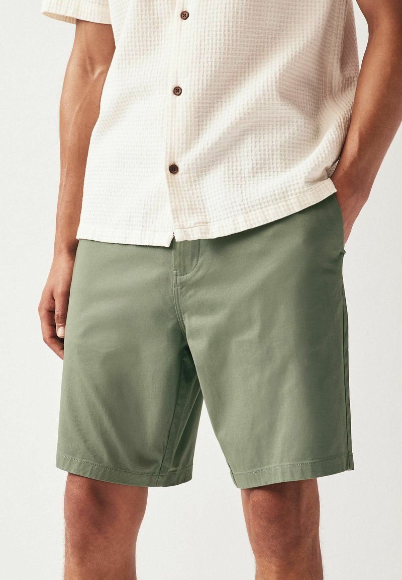 Shorts en coton verts avec poches latérales, coupe droite et une seule couture frontale. Portés avec une chemise à manches courtes texturée de couleur claire.