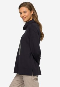 Pullover nero con collo alto, fessure laterali e texture morbida. Caratterizzato da una stampa grafica e vestibilità casual. Abbinato a pantaloni chiari.
