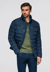 Marinblå puffjacka med horisontell quiltning, dragkedjefickor på bröstet och ståkrage, bärs över en grön ribbad tröja och blå jeans.