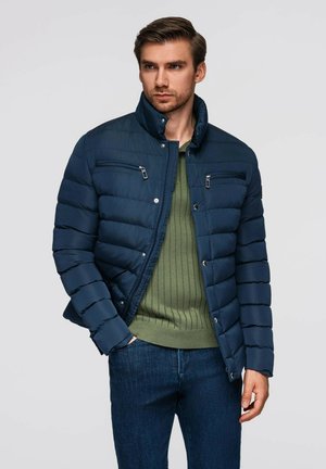 Giacca puffer blu navy con trapuntatura orizzontale, tasche sul petto con zip e colletto rialzato, indossata sopra un maglione verde a coste e jeans blu.