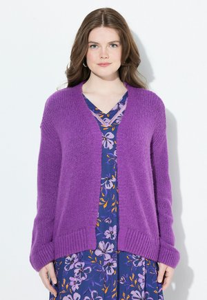 Frau mit braunen Haaren, die eine lila gestrickte Strickjacke über einem blauen Blumenkleid trägt, steht vor einem weißen Hintergrund.