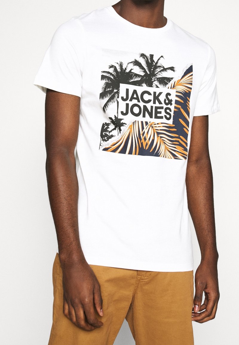 Personne portant un t-shirt blanc avec un graphisme de palmier et de feuille et le texte "JACK & JONES", associé à un pantalon marron clair.