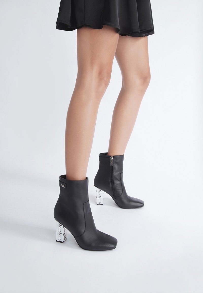 Jambes portant des bottines noires à talons blocs transparents, vêtues d'une jupe noire, debout sur un sol blanc, sur un fond clair.