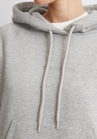 Sudadera gris claro hecha de un tejido suave, con un bolsillo tipo canguro, cordones y un discreto logo en relieve en el pecho.