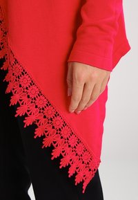 Červený top s dlouhými rukávy a krajkovým lemem s květinovými a listovými vzory. Detailní záběr na ruku spočívající na látce, která ukazuje texturu a detaily.