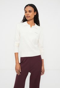 Kremfarget langermet polo-genser med ribbestrikket krage og nederkant, med en liten logo. Brukt med maroon løstsittende bukser.