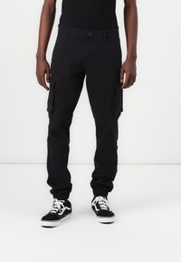 Schwarze Cargo-Hose mit tailliertem Bund, elastischen Bündchen und zwei großen Seitentaschen. Kombiniert mit schwarzen und weißen Turnschuhen.