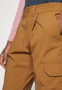 Brown cargo pants made of lightweight fabric featuring pleats, a side pocket, and a button closure at the waistband.