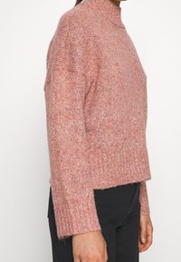 Pull rose texturé avec un col montant et des poignets côtelés. Coupe courte et carrée avec un ourlet légèrement raccourci. Tissu tricoté décontracté.