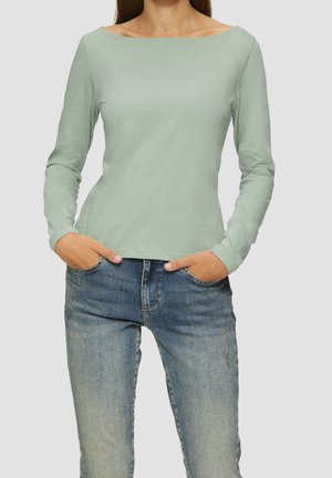 Femme portant une chemise à manches longues vert clair et un jean bleu, les mains dans les poches, sur un fond uni.