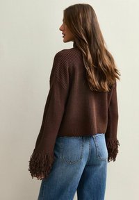 Pull en tricot marron avec des manches ouvertes et des accents en frange, associé à un jean en denim bleu clair, vu de dos.