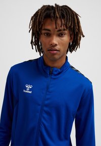 Blauer Sportjacke mit Reißverschluss aus strukturiertem Stoff, mit schwarzem Akzent auf den Schultern und einem weißen Logo auf der linken Brust.